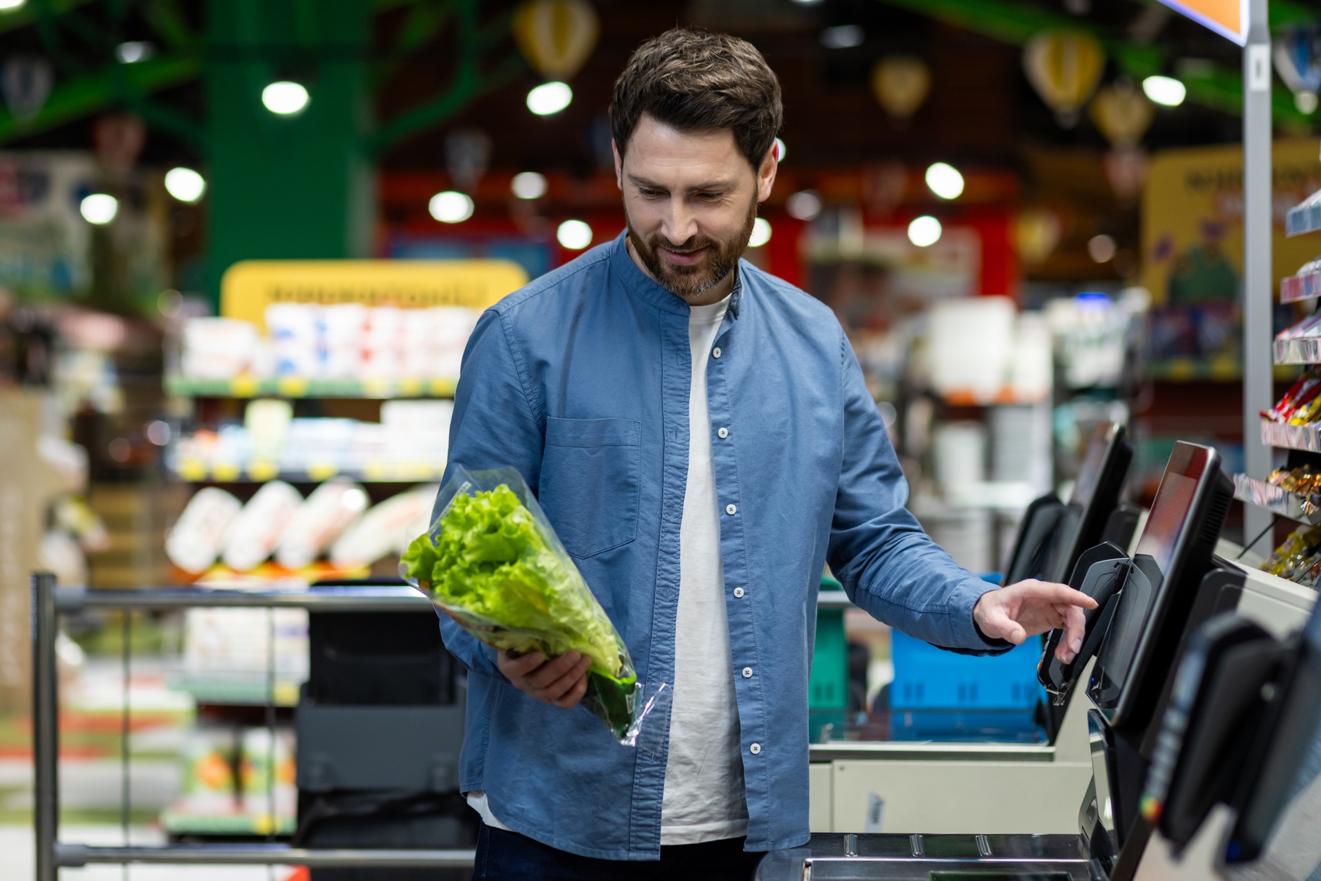 Man using self-checkout at grocery store while holding fresh lettuce, exemplifying modern shopping experience and convenience